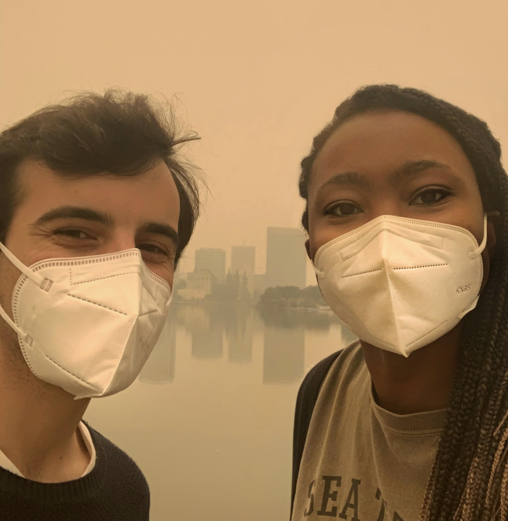 two volunteers, a latino middle eastern man and a Nigerian-American woman stand wearing white face masks with a smile in their eyes. Lake Merritt is in background, with downtown Oakland's buildings reflected, the sky is a disturbing brownish orange from wildfire smoke.